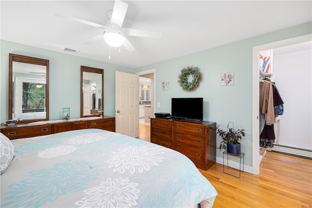 bedroom with baseboard heating, connected bathroom, ceiling fan, a walk in closet, and light hardwood / wood-style flooring