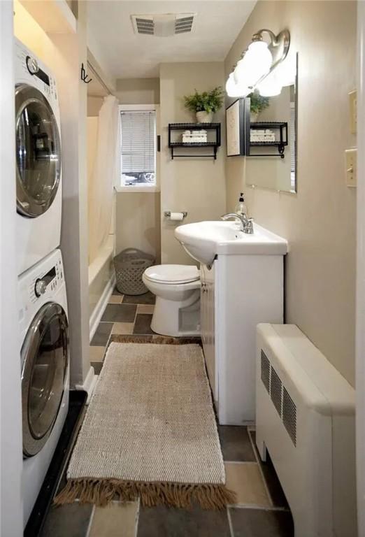 full bathroom featuring stacked washer / drying machine, vanity, toilet, shower / tub combo, and radiator heating unit