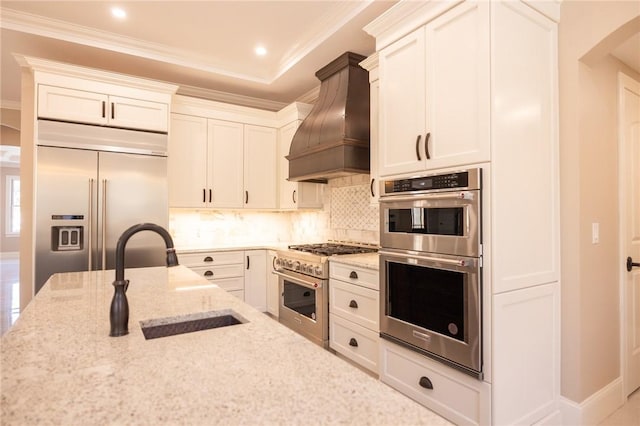 kitchen with light stone countertops, premium appliances, white cabinetry, premium range hood, and sink