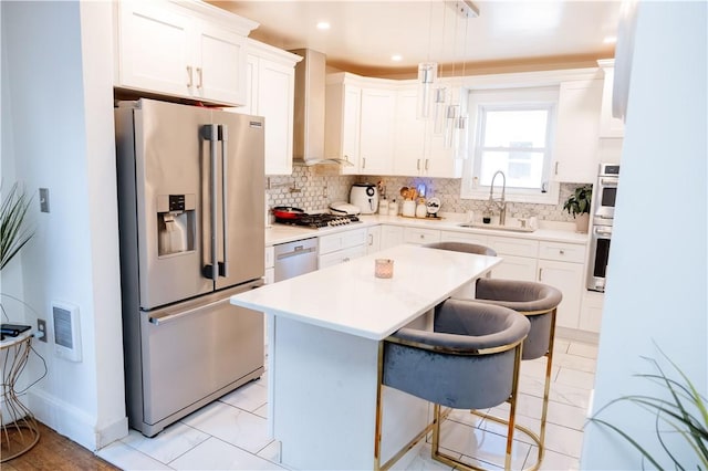 kitchen with white cabinets, wall chimney range hood, stainless steel appliances, sink, and hanging light fixtures