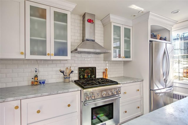 kitchen featuring light stone counters, wall chimney range hood, white cabinets, stainless steel appliances, and backsplash