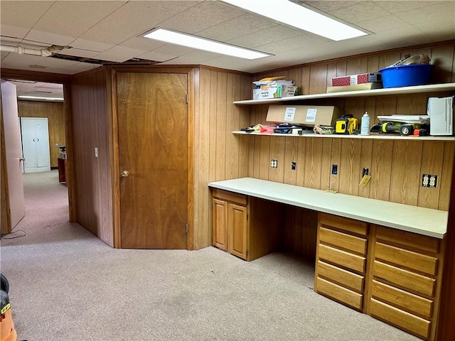 unfurnished office featuring a paneled ceiling, wooden walls, built in desk, and light carpet