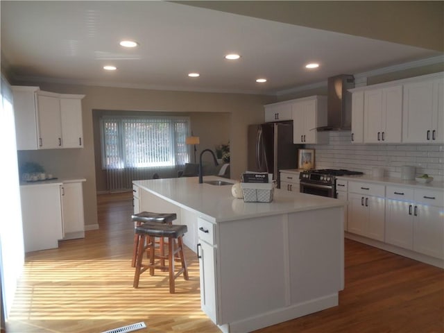 kitchen with sink, white cabinetry, a center island with sink, stainless steel appliances, and wall chimney range hood