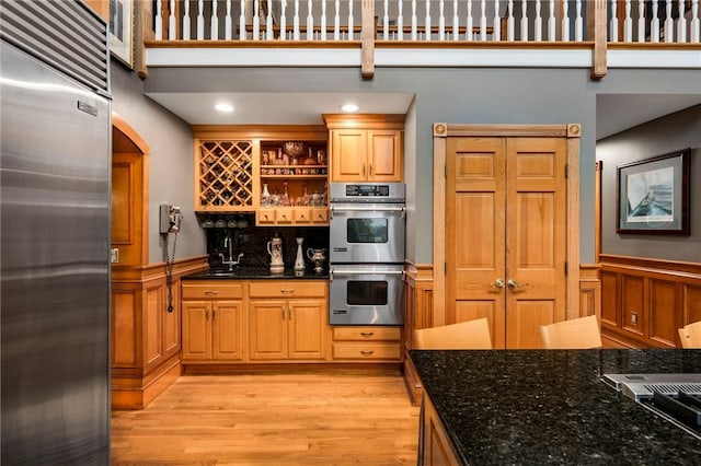 kitchen featuring open shelves, appliances with stainless steel finishes, light wood-style floors, wainscoting, and dark stone countertops