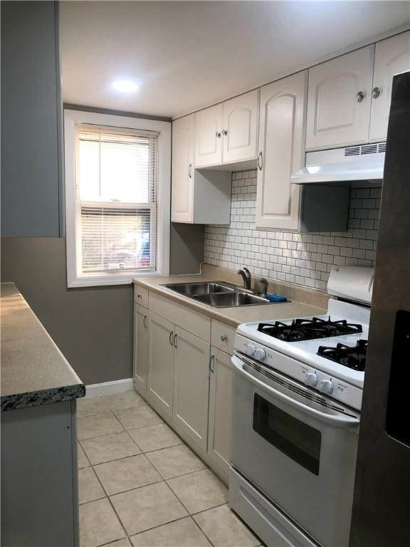 kitchen with light tile patterned floors, sink, backsplash, white cabinets, and white gas range