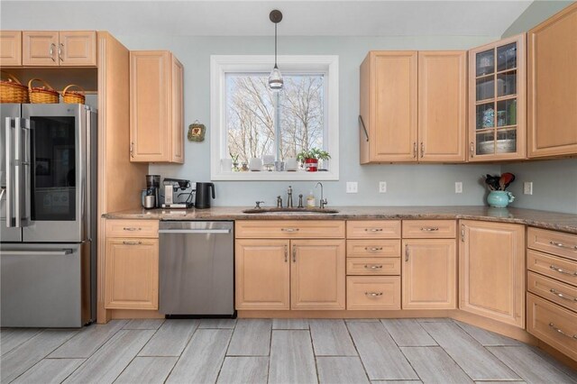 kitchen with glass insert cabinets, light brown cabinetry, decorative light fixtures, stainless steel appliances, and a sink