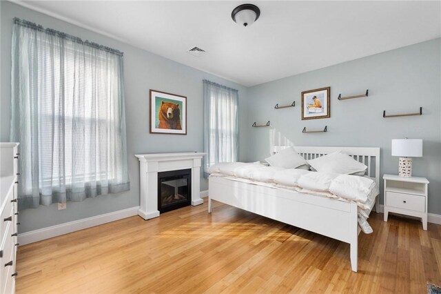 bedroom with a glass covered fireplace, visible vents, light wood-style floors, and baseboards