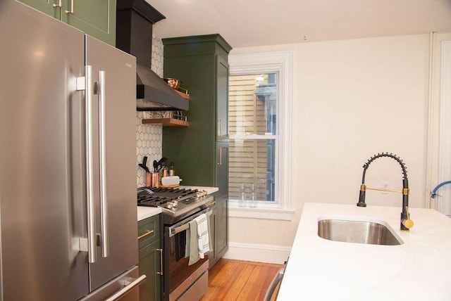 kitchen with sink, green cabinets, stainless steel appliances, wall chimney range hood, and light hardwood / wood-style flooring