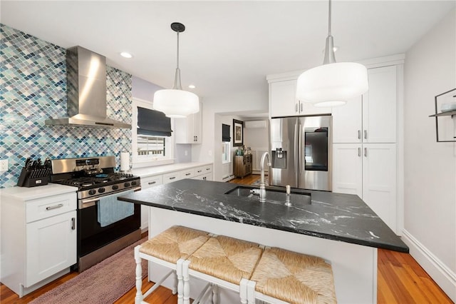 kitchen featuring appliances with stainless steel finishes, wall chimney exhaust hood, an island with sink, and hanging light fixtures