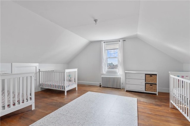 bedroom featuring a crib, baseboards, radiator heating unit, wood finished floors, and vaulted ceiling