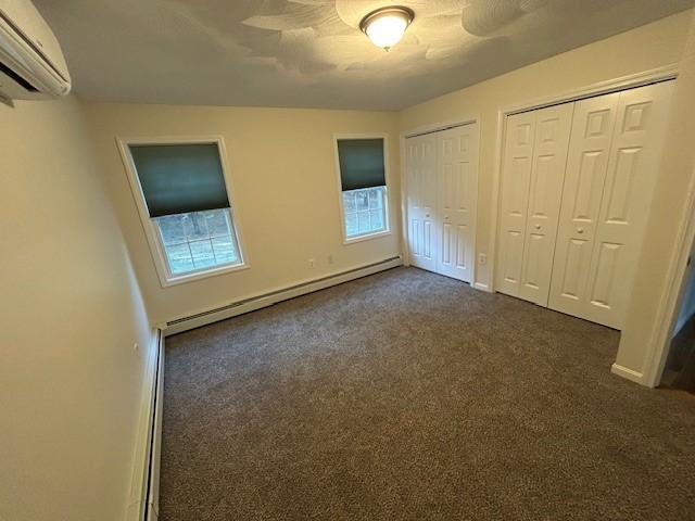 unfurnished bedroom with a baseboard heating unit, dark carpet, multiple closets, and a wall mounted AC