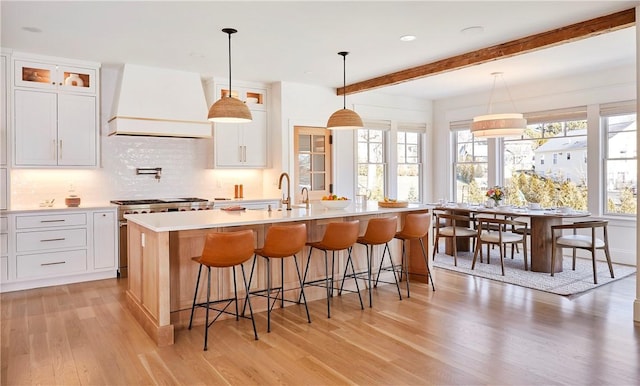 kitchen with hanging light fixtures, high end stove, an island with sink, white cabinets, and custom exhaust hood