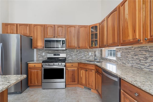 kitchen with light stone counters, brown cabinets, glass insert cabinets, stainless steel appliances, and a sink