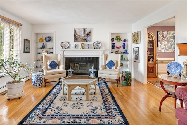 living room featuring a brick fireplace, built in shelves, wood-type flooring, and baseboard heating