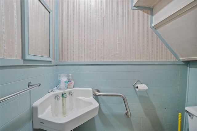 half bathroom with toilet, a wainscoted wall, a sink, tile walls, and wallpapered walls