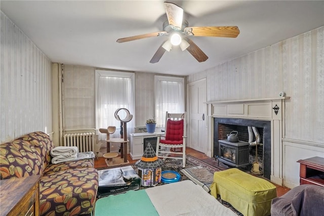 living room featuring wallpapered walls, a ceiling fan, a wood stove, and radiator