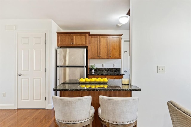 kitchen featuring a kitchen bar, brown cabinets, wood finished floors, freestanding refrigerator, and dark stone counters