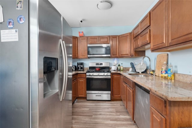 kitchen featuring light wood-style flooring, appliances with stainless steel finishes, brown cabinetry, a sink, and light stone countertops