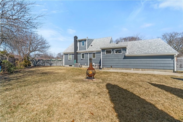 rear view of house featuring a lawn, fence, a fire pit, roof with shingles, and a chimney