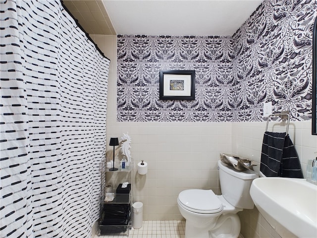 full bathroom featuring toilet, a wainscoted wall, a sink, tile walls, and wallpapered walls