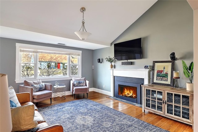 living area with baseboards, visible vents, a fireplace with flush hearth, wood finished floors, and vaulted ceiling