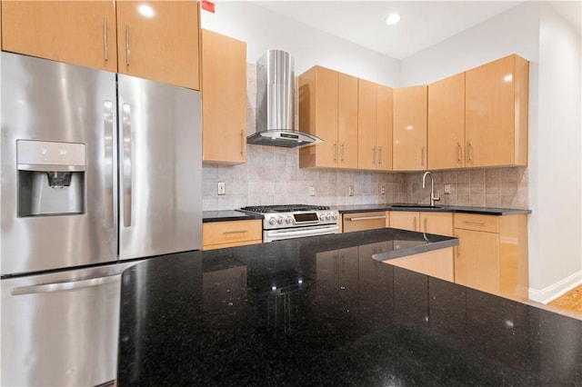 kitchen with light brown cabinetry, appliances with stainless steel finishes, a sink, wall chimney range hood, and dark stone countertops