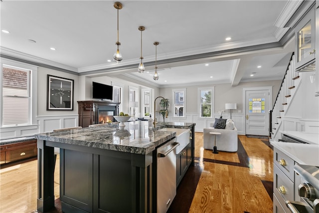 kitchen featuring a center island with sink, dark stone counters, dishwasher, a glass covered fireplace, and hanging light fixtures