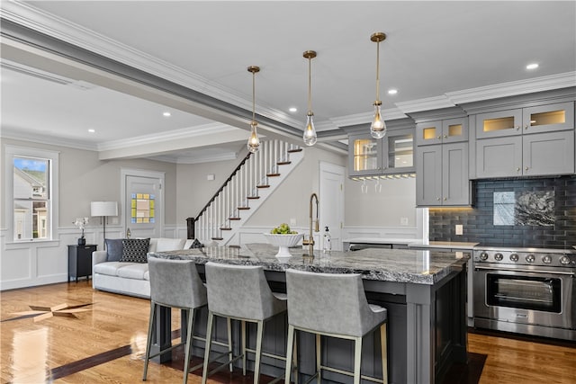 kitchen featuring a center island with sink, gray cabinets, high end stainless steel range oven, glass insert cabinets, and dark stone counters