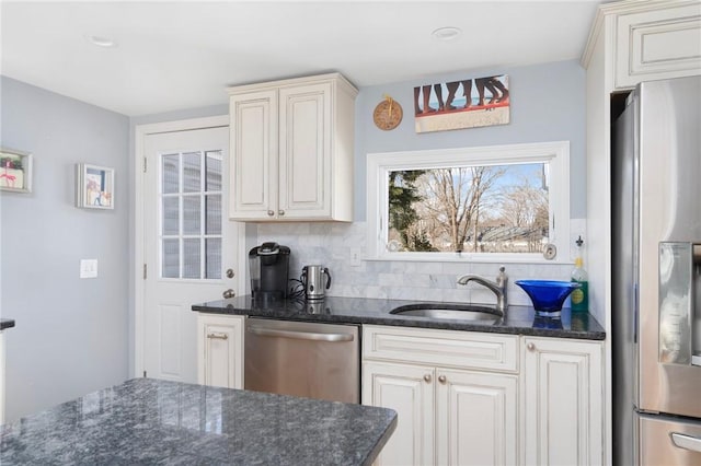 kitchen with cream cabinetry, stainless steel appliances, backsplash, a sink, and dark stone countertops