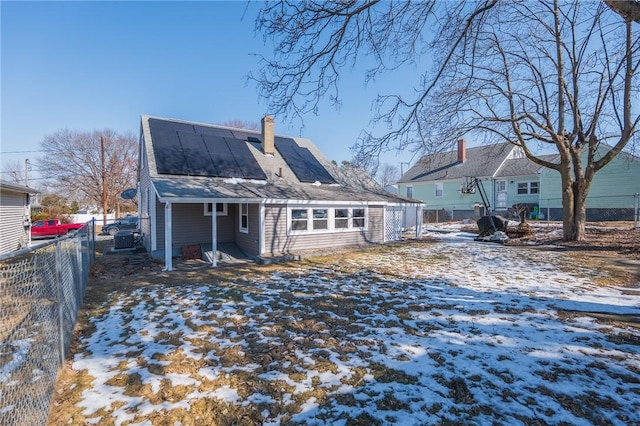 snow covered house featuring a chimney, fence, and solar panels