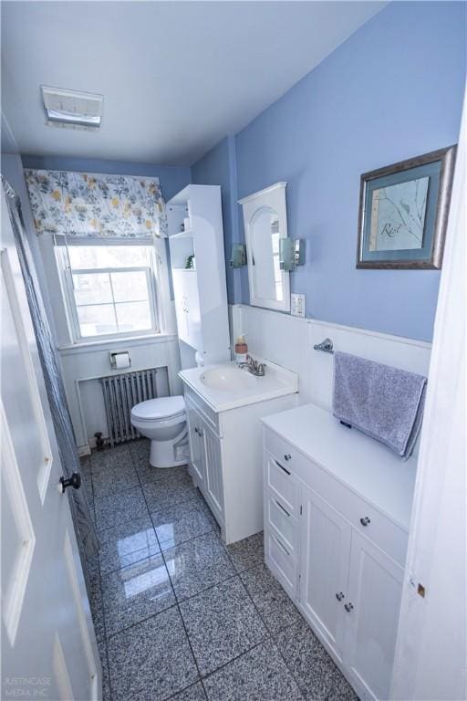 bathroom with toilet, granite finish floor, vanity, wainscoting, and radiator heating unit