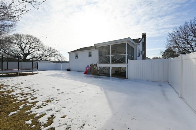 snow covered rear of property featuring a sunroom, a fenced backyard, a trampoline, and a chimney