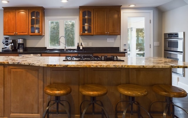 kitchen with double oven, a sink, gas stovetop, brown cabinets, and dark stone counters