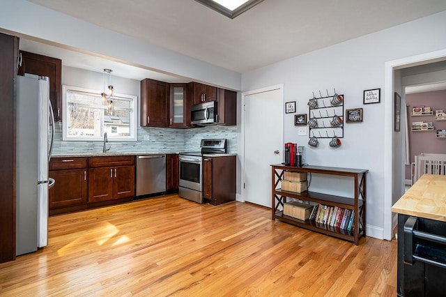 kitchen with hanging light fixtures, decorative backsplash, appliances with stainless steel finishes, light wood-style floors, and glass insert cabinets