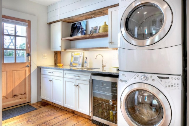 clothes washing area featuring light wood finished floors, cabinet space, a sink, wine cooler, and stacked washer / drying machine