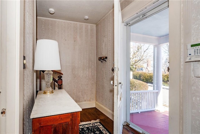 doorway to outside with dark wood-type flooring, a textured ceiling, baseboards, and wallpapered walls