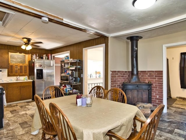 dining space featuring wood walls, a wood stove, ceiling fan, stone finish flooring, and wainscoting