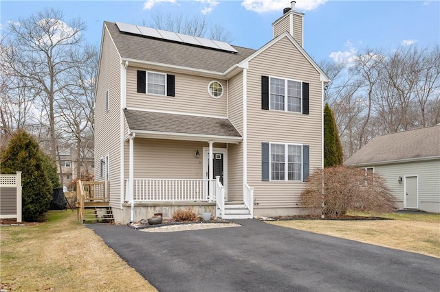 traditional home with a shingled roof, a chimney, roof mounted solar panels, a front lawn, and a porch