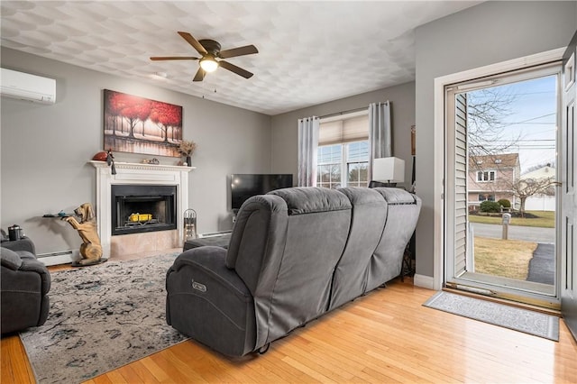 living room featuring a wall unit AC, light wood-style floors, a baseboard heating unit, a fireplace with flush hearth, and a ceiling fan