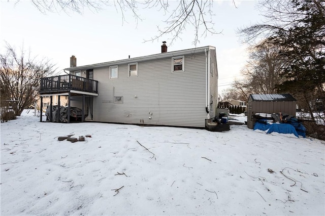 snow covered house with a shed, a chimney, a wooden deck, and an outbuilding