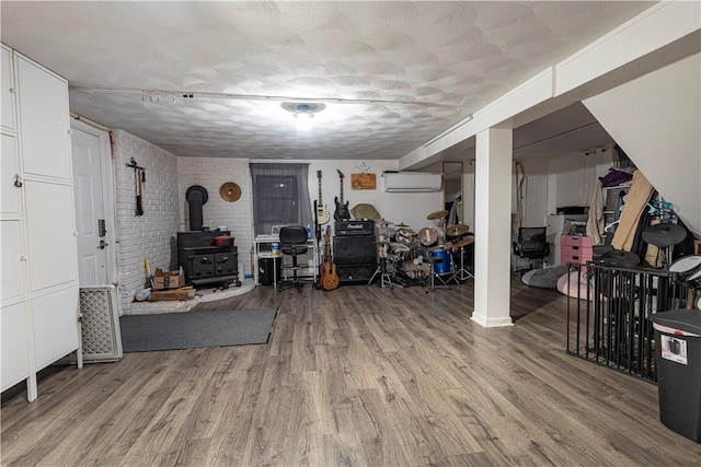 finished basement with a wall unit AC, light wood-style flooring, a wood stove, a textured ceiling, and brick wall