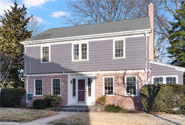 colonial-style house featuring brick siding, a chimney, and roof with shingles