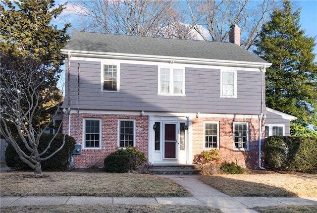 colonial home with roof with shingles, a chimney, and brick siding