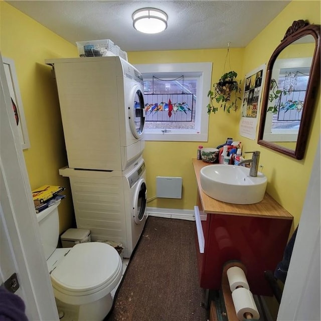 half bathroom featuring toilet, vanity, stacked washing maching and dryer, a textured ceiling, and baseboards