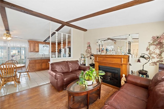 living room featuring vaulted ceiling with beams, ceiling fan, light wood finished floors, and a glass covered fireplace