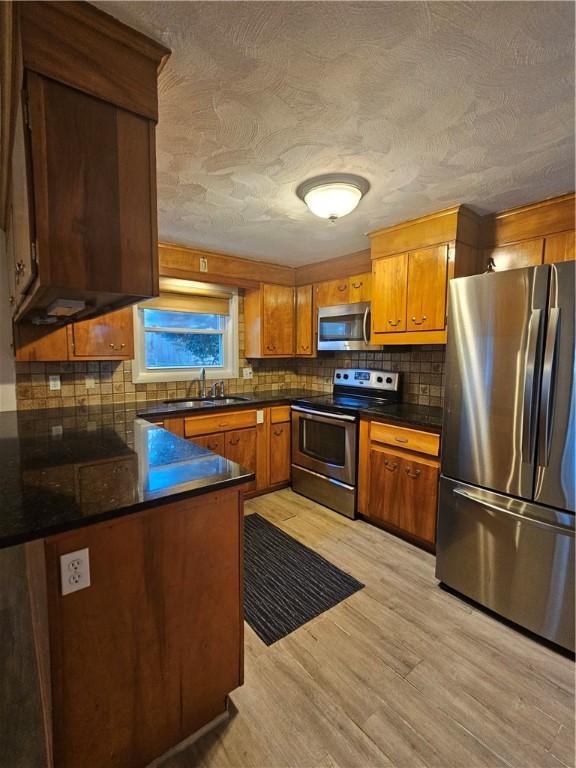 kitchen with a sink, appliances with stainless steel finishes, light wood finished floors, brown cabinetry, and dark countertops