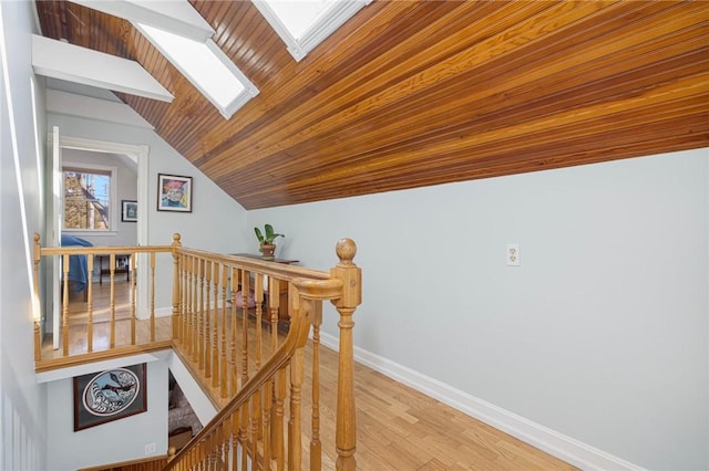 corridor featuring vaulted ceiling with skylight, wooden ceiling, an upstairs landing, baseboards, and light wood finished floors