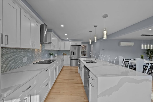 kitchen with a wall unit AC, appliances with stainless steel finishes, wall chimney range hood, white cabinetry, and a sink