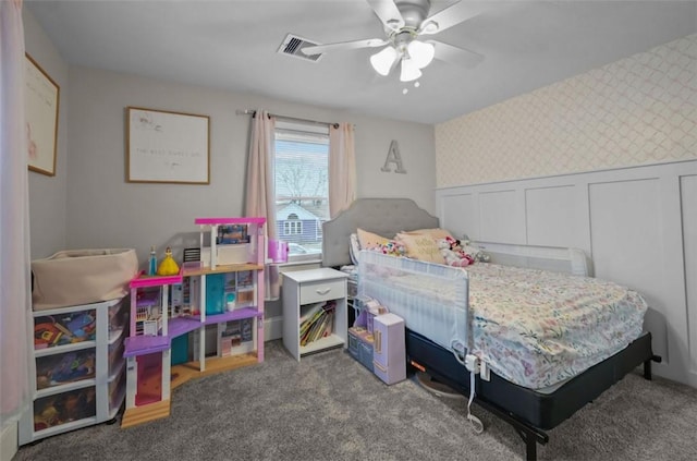 carpeted bedroom with a decorative wall, a ceiling fan, visible vents, wainscoting, and wallpapered walls