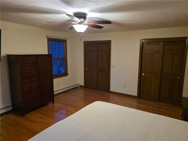 unfurnished bedroom featuring a baseboard heating unit, wood finished floors, a ceiling fan, baseboards, and multiple closets